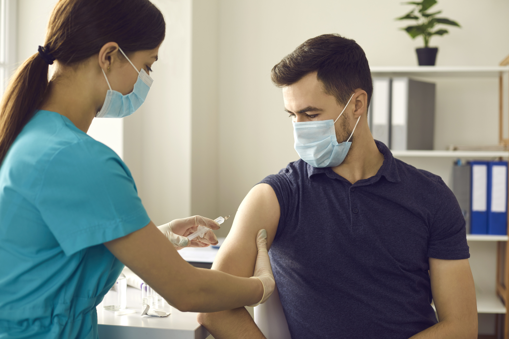 Professional nurse in medical face mask giving flu shot to male patient during seasonal vaccination campaign. Doctor injecting young man with influenza, AIDS or Covid-19 vaccine at clinic office
